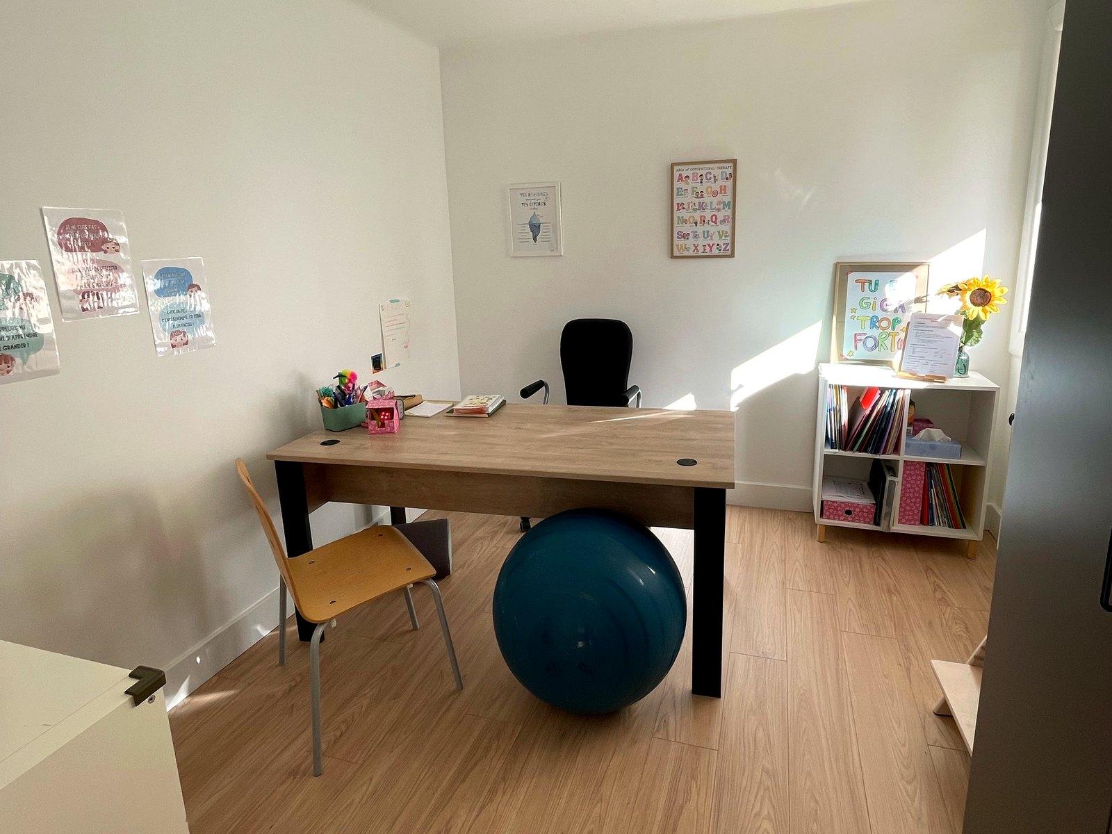 Un bureau du cabinet : table de travail, ballon d'assise, chaise enfant et bibliothèque de jeux.