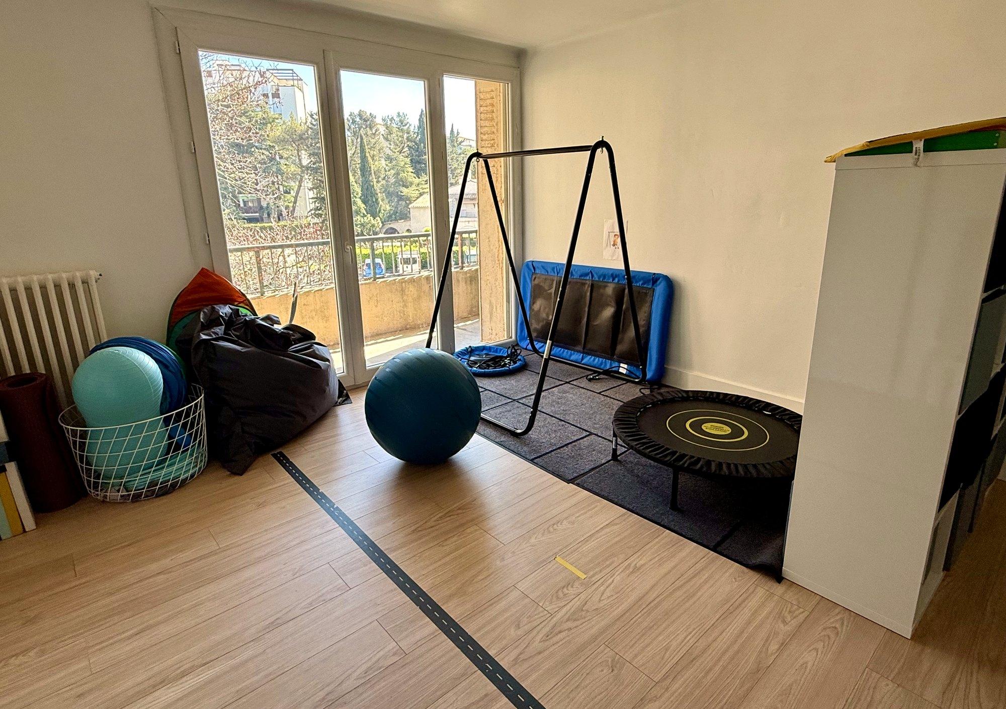 La salle de motricité : ballons, trampoline, balançoire et tapis, devant la baie vitrée.
